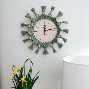 May include: A round wall clock featuring a sage green macrame border with decorative tassels and light wooden beads. The clock face is off-white with black ornate hands and thin black markers. The text "Vano" is printed on the clock face.