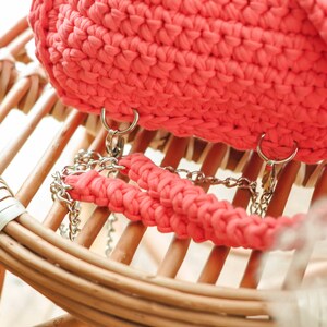 May include: A coral-colored, crocheted handbag with silver chain straps. The bag is sitting on a light brown wicker chair. The bag's texture is visible, and the chain straps are attached with silver rings.