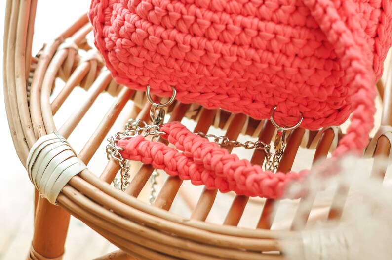 May include: A coral-colored, crocheted handbag with silver chain straps. The bag is sitting on a light brown wicker chair. The bag's texture is visible, and the chain straps are attached with silver rings.