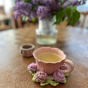 May include: A pink teacup filled with coffee sits on a crocheted coaster with pink and purple flower accents. A small, brown cup is behind the teacup. A vase of purple flowers is in the background.