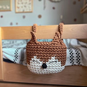Può includere: Un cesto appeso in crochet marrone e bianco a forma di volpe con occhi neri e naso nero. Il cesto è appeso a uno scaffale di legno.