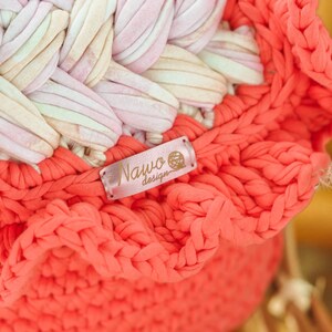 May include: Close-up of a handmade coral-colored crochet basket with a ruffled edge. The basket features a woven design in shades of pink, white, and beige. A small label with the text "Nawo design" is attached to the basket.