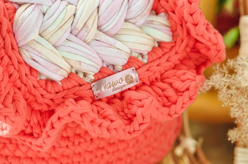 May include: Close-up of a handmade coral-colored crochet basket with a ruffled edge. The basket features a woven design in shades of pink, white, and beige. A small label with the text "Nawo design" is attached to the basket.