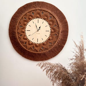 May include: A brown macrame wall clock with a wooden face and black hands. The clock is surrounded by a fringe of brown yarn.