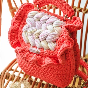 May include: Coral-colored crochet handbag with a woven center panel of pink and white fabric. The bag features a rounded shape, a handle, and a shoulder strap. A small tag with the text "Nawo design" is visible.