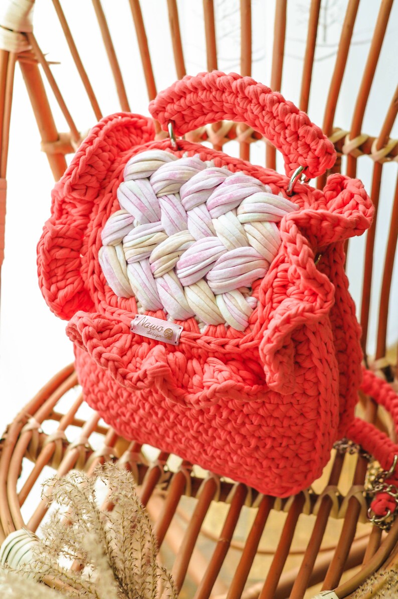 May include: Coral-colored crochet handbag with a woven center panel of pink and white fabric. The bag features a rounded shape, a handle, and a shoulder strap. A small tag with the text "Nawo design" is visible.