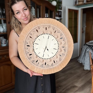 May include: A woman holds a large, round, beige macrame wall clock with a wooden clock face. The clock has a fringe border and a detailed mandala design.