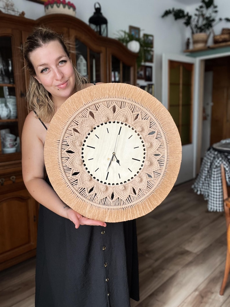 May include: A woman holds a large, round, beige macrame wall clock with a wooden clock face. The clock has a fringe border and a detailed mandala design.