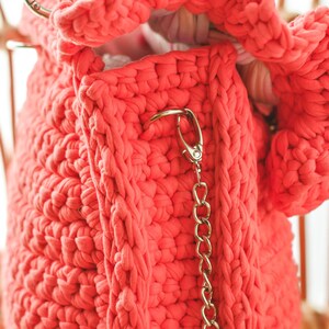 May include: A coral-colored, chunky knit bag with a gold-toned chain strap and clasp. The bag is made of thick, textured yarn, and the interior is visible. The bag is hanging on a light-colored wooden chair.