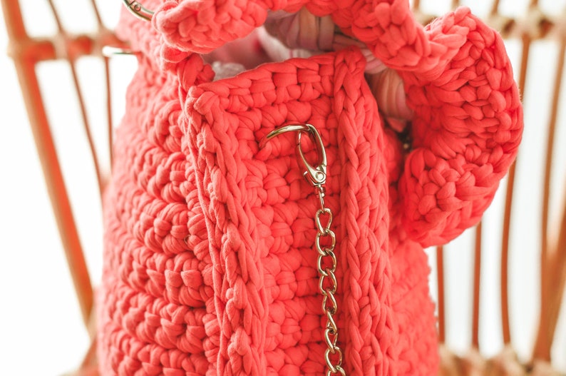 May include: A coral-colored, chunky knit bag with a gold-toned chain strap and clasp. The bag is made of thick, textured yarn, and the interior is visible. The bag is hanging on a light-colored wooden chair.