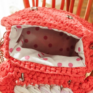 May include: Coral-colored, handmade crochet bag with a silver chain strap. The bag's interior features a white fabric with pink polka dots. The bag is open, revealing the interior. The bag is sitting on a wooden chair.