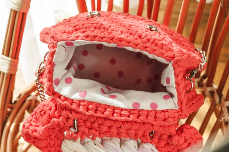 May include: Coral-colored, handmade crochet bag with a silver chain strap. The bag's interior features a white fabric with pink polka dots. The bag is open, revealing the interior. The bag is sitting on a wooden chair.