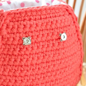 May include: A coral-colored, crocheted handbag with a white polka-dot lining. The bag features two silver magnetic closures and a silver chain. The texture of the crochet is visible, and the bag appears to be handmade.