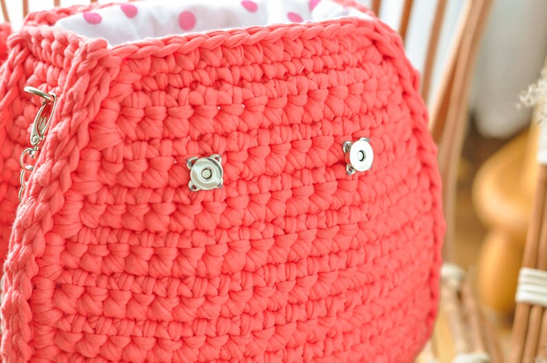May include: A coral-colored, crocheted handbag with a white polka-dot lining. The bag features two silver magnetic closures and a silver chain. The texture of the crochet is visible, and the bag appears to be handmade.