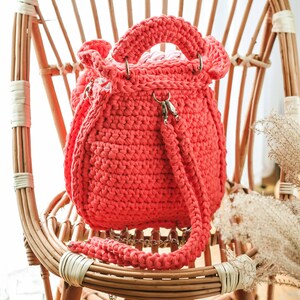 May include: A coral-colored, handmade crochet bag with a drawstring closure and a shoulder strap. The bag features a rounded shape, metal hardware, and a sturdy handle. It is displayed on a natural wicker chair.