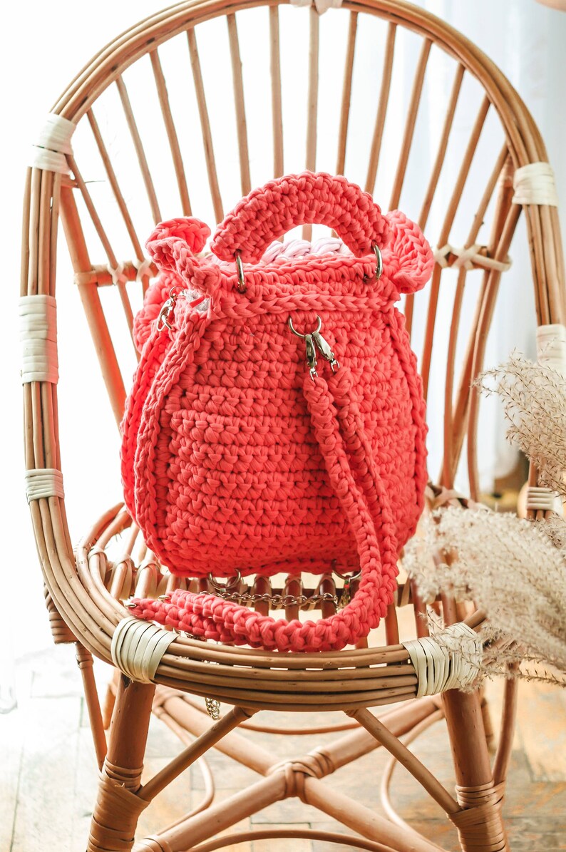 May include: A coral-colored, handmade crochet bag with a drawstring closure and a shoulder strap. The bag features a rounded shape, metal hardware, and a sturdy handle. It is displayed on a natural wicker chair.