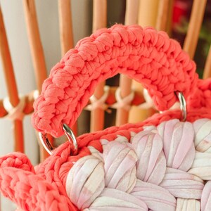 May include: Close-up of a handmade coral-colored crochet bag with a woven design in shades of pink and white. The bag features a thick, rounded handle attached with silver metal rings. The background shows a wooden chair.