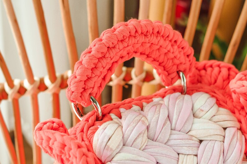 May include: Close-up of a handmade coral-colored crochet bag with a woven design in shades of pink and white. The bag features a thick, rounded handle attached with silver metal rings. The background shows a wooden chair.