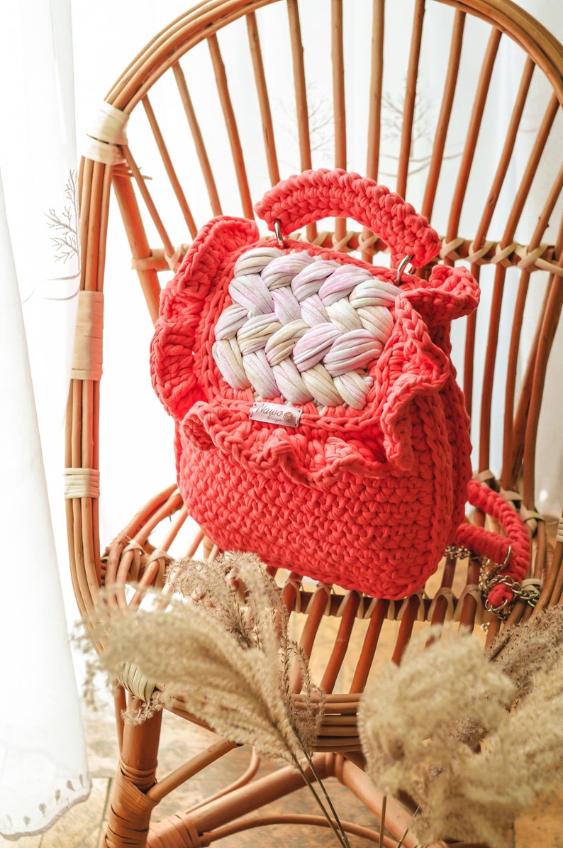 May include: Coral-colored crochet handbag with a woven center panel of white and pink. The bag has a ruffled edge and a matching handle. It is sitting on a wicker chair.