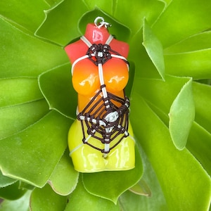 May include: A colorful candle shaped like a torso, featuring a gradient of red, orange, and yellow. The candle is adorned with a macrame design in brown and white, and is suspended by a silver loop. The background is a vibrant green succulent.
