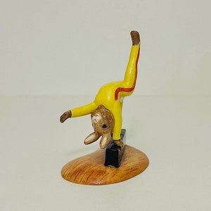 Puede incluir: Una pequeña figurita de conejo marrón vestida con un atuendo amarillo y rojo está haciendo una parada de manos sobre un bloque negro. La figurita está sobre una base de madera.