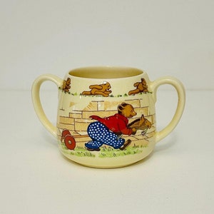 May include: A vintage ceramic cup with two handles, featuring a whimsical illustration of a bear pulling a toy horse, surrounded by other bears. The cup is cream-colored with brown, red, blue, and yellow accents.