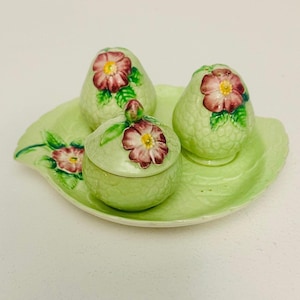Puede incluir: Un juego de condimentos de cerámica vintage. Cuenta con una bandeja en forma de hoja verde claro, dos saleros y pimenteros en forma de pera y un pequeño tarro con tapa. Cada pieza está adornada con un diseño floral en rosa y verde.