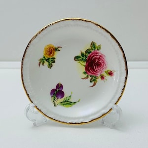 May include: A white porcelain plate with a gold rim and floral design. The plate features a pink rose, a yellow rose, and purple berries.