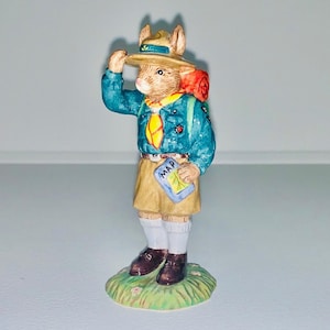 May include: A ceramic figurine of a rabbit wearing a scout uniform, hat, and backpack. The rabbit is holding a map that says "MAP".