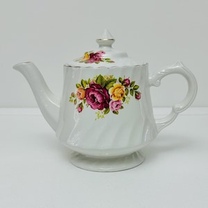Może przedstawiać: Biały porcelanowy czajnik z kwiatowym wzorem przedstawiającym różowe, żółte i czerwone róże. Czajnik ma żebrowane ciało i pozłacaną pokrywkę.