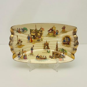 May include: A white ceramic serving dish with a gold rim and a decorative design of English landmarks. The dish features illustrations of buildings, people, and scenes from different cities in England.