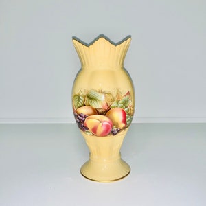 May include: A yellow ceramic vase with a fruit design. The vase has a fluted body and a scalloped rim. The fruit design features peaches, grapes, and leaves.