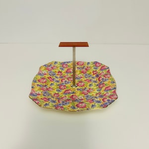 May include: A yellow floral cake stand with a brown wooden top and a silver metal stand.