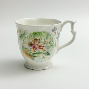 May include: A white ceramic mug with a scalloped rim and a decorative handle. The exterior features a colourful illustration of a frog wearing a pink jacket and carrying a basket, standing on a lily pad in a pond with green reeds and white flowers.