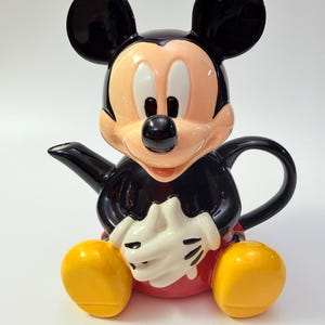 May include: A ceramic teapot shaped like Mickey Mouse, featuring his iconic black ears, peach-coloured face, white gloves, red shorts, and yellow shoes. The teapot has a black spout extending from the side and a black handle on the back.