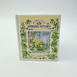 Puede incluir: Un adorno de cerámica con forma de libro que presenta la ilustración Spring Story de Brambly Hedge de Jill Barklem. El frente muestra un borde amarillo, follaje verde y una escena central de flores amarillas y un ratón pequeño, con el texto Spring Story.