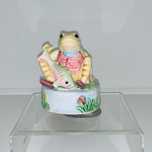 Peut inclure: Un carillon en porcelaine représentant une grenouille tenant un poisson. La grenouille porte un gilet rose et bleu et a des rayures jaunes sur les pattes. Le carillon est décoré d'herbe verte et d'un étang bleu.
