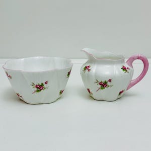 Może przedstawiać: Biały zestaw cukiernicy i mlecznika z różowym wzorem kwiatowym. Cukiernica ma falisty brzeg, a mlecznik ma różowy uchwyt.