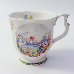 May include: A white ceramic mug featuring a colorful illustration of anthropomorphic animals in a boat on a river. The scene includes flowers, reeds, and dragonflies. The mug has a scalloped rim, a decorative handle, and a green painted base trim.