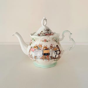 Può includere: Una teiera in ceramica bianca con coperchio, con un'illustrazione stravagante di topi a tavola. La teiera ha un beccuccio e un manico curvi, con una base verde chiaro e dettagli floreali. Il coperchio presenta la scritta "Brambly Hedge".