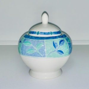 Op de afbeelding: Een witte suikerpot van keramiek met een deksel. De pot heeft een blauw en groen bloemenpatroon op een witte achtergrond. Het deksel heeft een blauw en wit patroon.