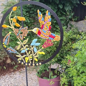 Peut inclure: Un motif de colibri et de fleurs en mosaïque dans un cercle en métal noir. Le colibri est composé de carreaux rouges, jaunes, bleus et oranges. L'œuvre est placée devant un fond de feuillage vert et un pot de fleurs rose.