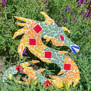 Peut inclure: Une sculpture de dragon en mosaïque colorée, ornée de carreaux jaunes, oranges, verts, rouges et bleus. Le dragon est placé dans un jardin avec un feuillage vert et des fleurs violettes. La mosaïque est faite de petits carreaux irréguliers.