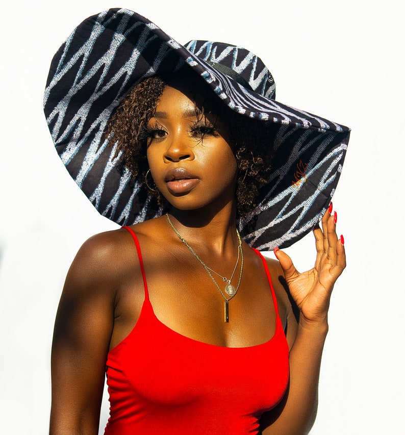 May include: A woman wearing a large, black and white patterned sun hat with a wide brim. She is wearing a red tank top and a silver necklace with a pendant.