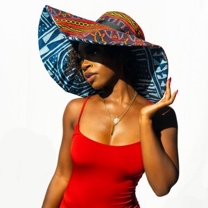 May include: A woman wearing a large, wide-brimmed sun hat with a colorful geometric pattern. The hat is blue, red, yellow, and black. She is wearing a red sleeveless dress and has a gold necklace with a pendant.