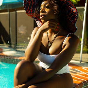May include: A woman wearing a white swimsuit and a large, colorful sun hat sits on the edge of a swimming pool. The hat has a blue, red, and yellow pattern.