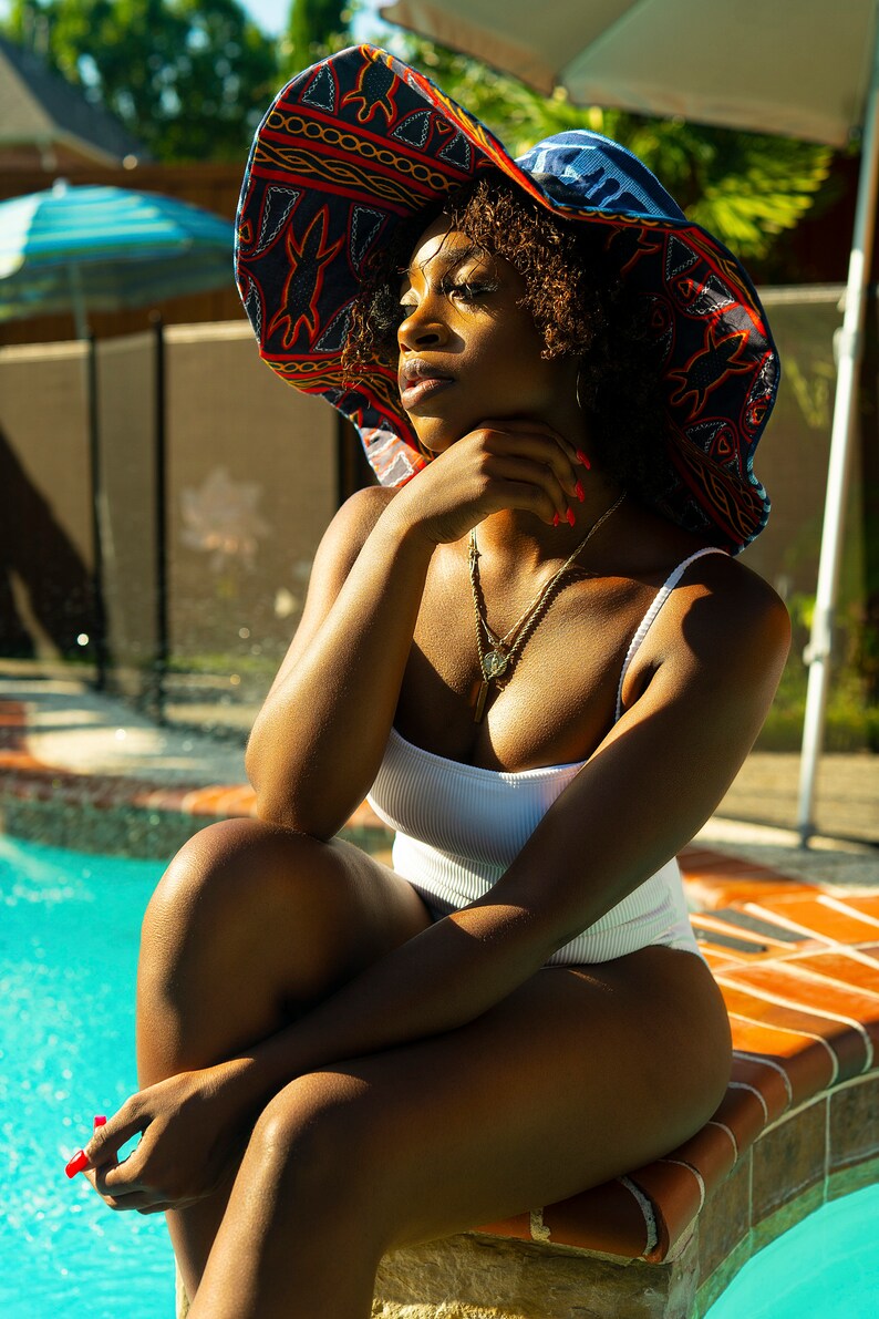 May include: A woman wearing a white swimsuit and a large, colorful sun hat sits on the edge of a swimming pool. The hat has a blue, red, and yellow pattern.