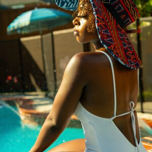 May include: A woman wearing a white swimsuit with a tie back sits by a pool. She is wearing a large, colorful sun hat with a geometric pattern.