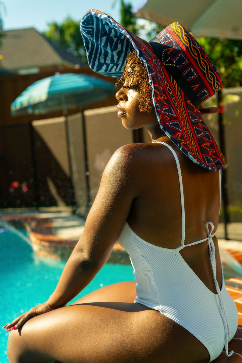 May include: A woman wearing a white swimsuit with a tie back sits by a pool. She is wearing a large, colorful sun hat with a geometric pattern.