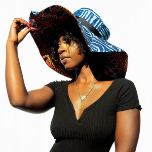 May include: A woman wearing a black shirt with a large, colorful sun hat with a blue and red patterned design. The hat has a wide brim and a black ribbon around the crown.